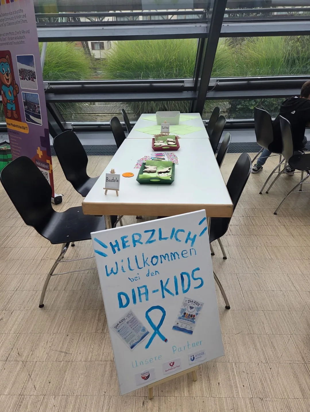 Tisch der Dia-Kids mit Willkommensschild, Flyern und Schälchen, im Hintergrund eine Fensterfront